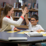 Woman tutor and young boy student sitting at a table and high fiving.