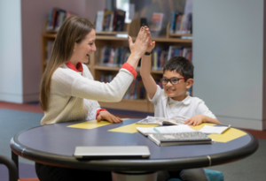 Woman tutor and young boy student sitting at a table and high fiving.